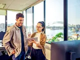 Two employees looking at a tablet, discussing work, with the Sydney Harbour Bridge seen through their office window